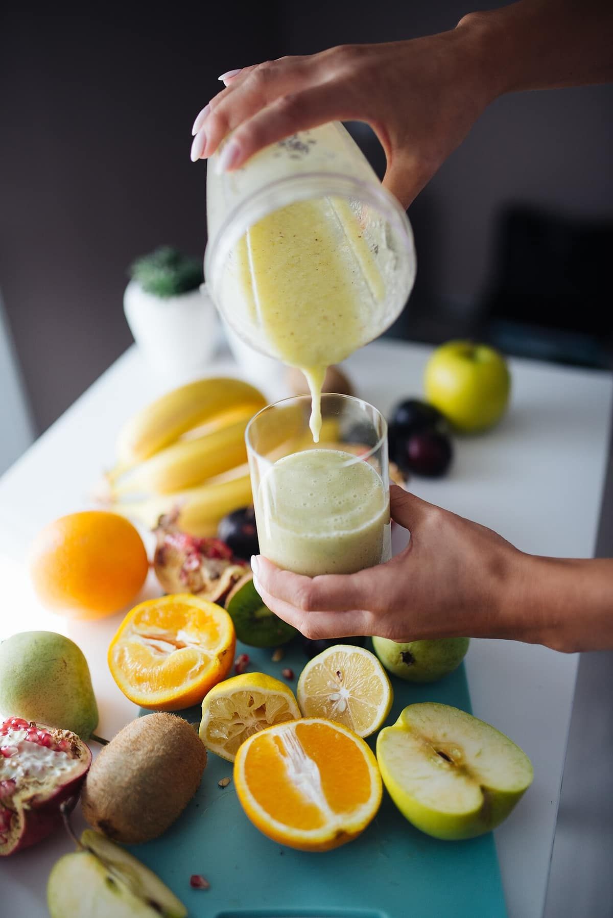 A person pours a pale fruit smoothie from a blender jar into a glass, surrounded by cut fruit