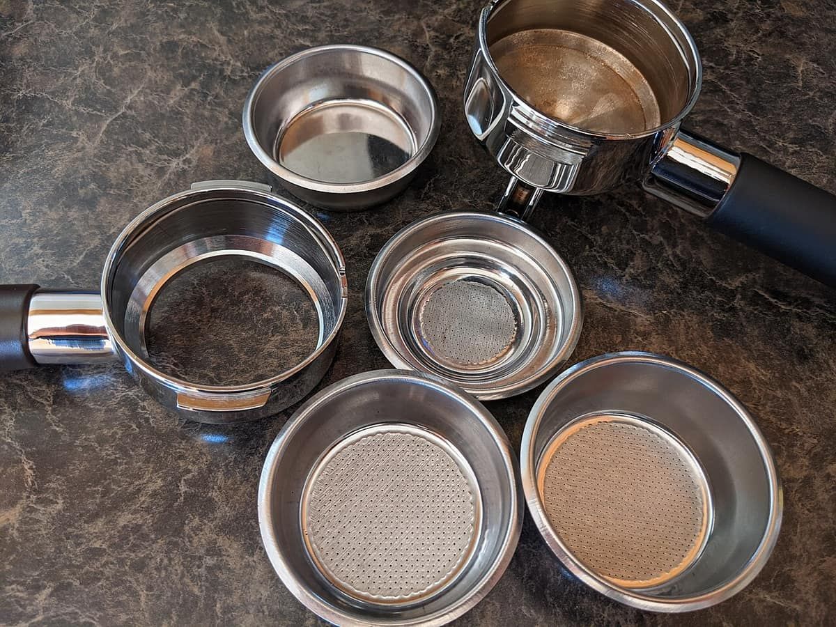 Two portafilters and several stainless filter baskets on a countertop