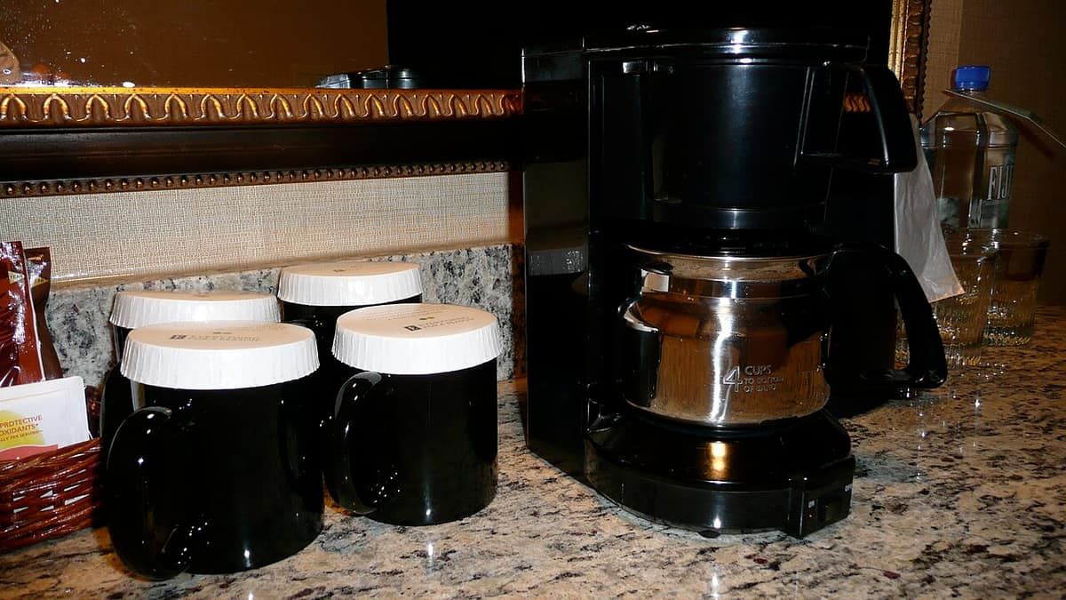 A drip coffee maker with a glass carafe on a hotel-room counter beside canisters of coffee and tea