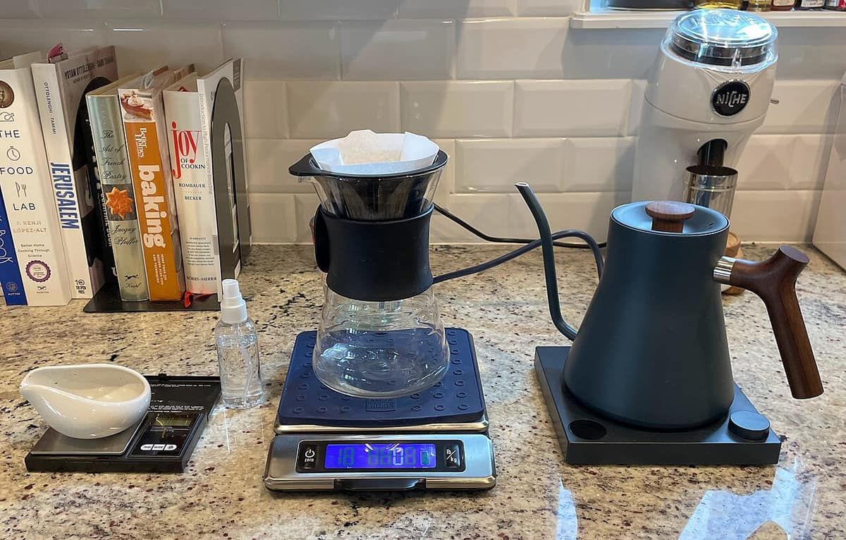 A pour-over coffee setup on a counter: a dripper on a digital scale, a gooseneck kettle and a grinder
