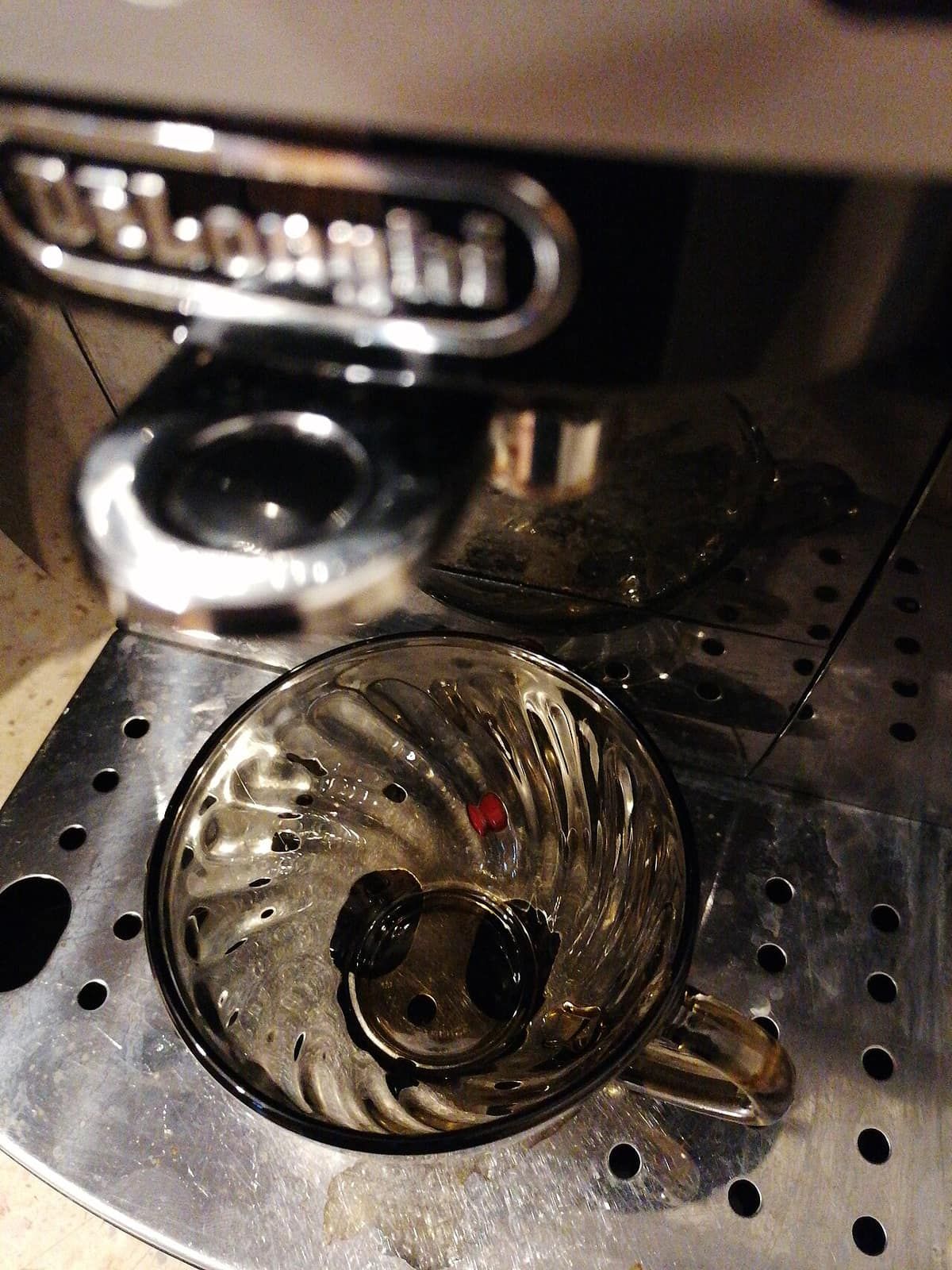 Close-up of a glass cup sitting on the drip tray under the group head of a De'Longhi espresso machine