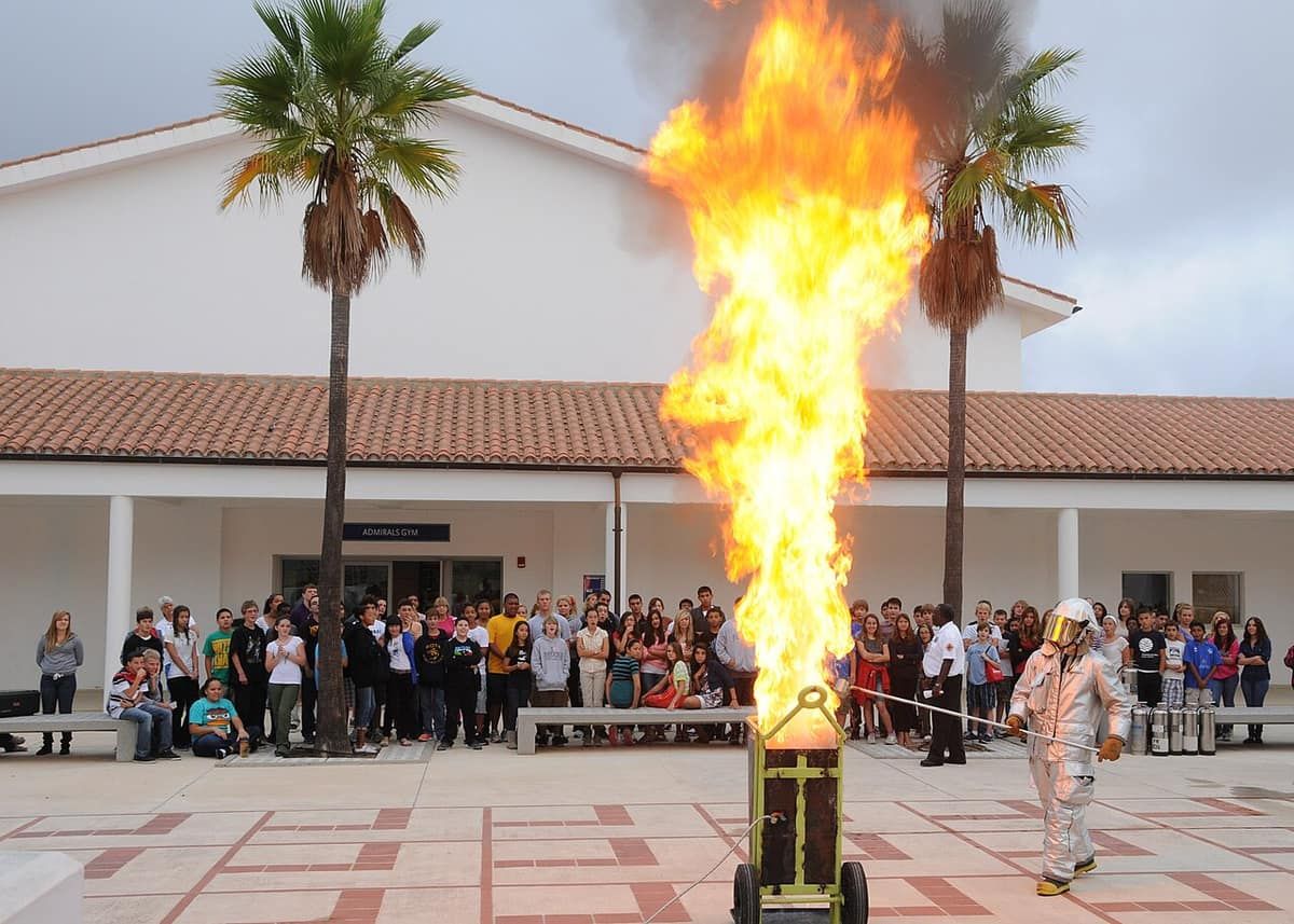 A tall fireball rises from a burning pot in an outdoor demonstration while a crowd of students watches from a distance