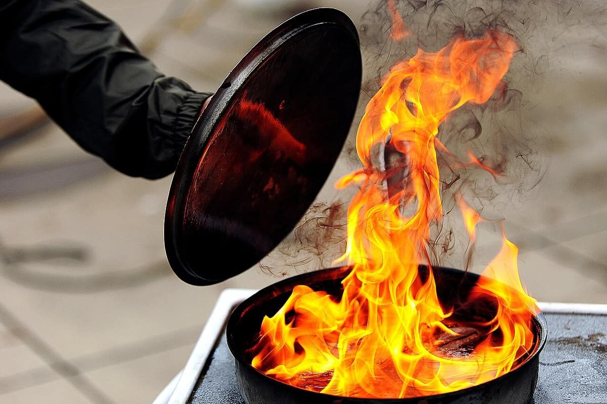 A person in a protective glove lowers a metal lid onto a pan of burning oil during a fire-safety demonstration