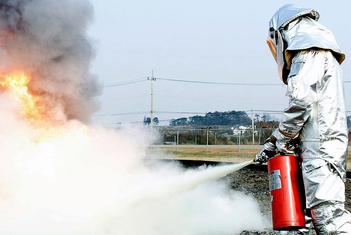 A firefighter in a silver proximity suit sprays a portable fire extinguisher at a training fire