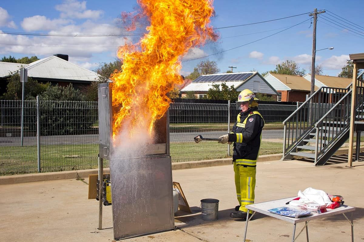 Firefighter demonstrating a burning cooking-oil fire in an outdoor training rig, with a tall fireball